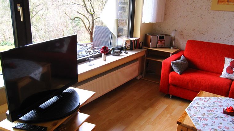 Ein gemütliches Wohnzimmer mit einem roten Sofa und einem Fernseher. Große Fenster lassen viel Tageslicht herein.