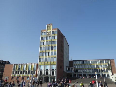 Een modern gebouw met een opvallende gevel en een hoge toren. Mensen staan ervoor en genieten van de zonnige dag.