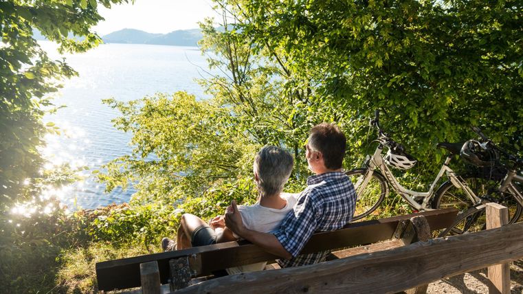 A couple is sitting on a bench, enjoying the view of the water and nature. Two bicycles are standing next to the bench.
