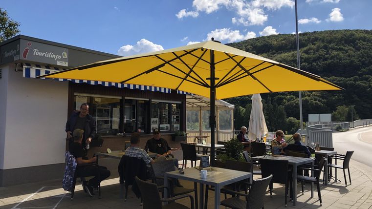 Een gezellig café buiten met een grote gele parasol. Gasten zitten aan tafels en genieten van de zon.