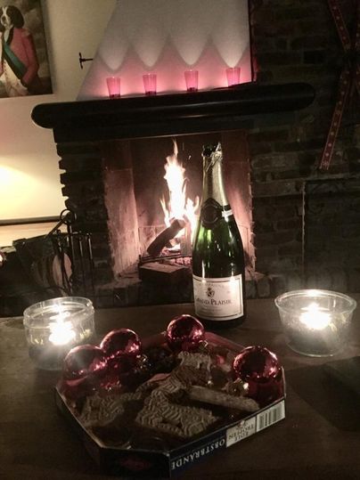 A cozy fireplace with a blazing fire and a glass of sparkling wine. On the table are gingerbread cookies and red Christmas ornaments.