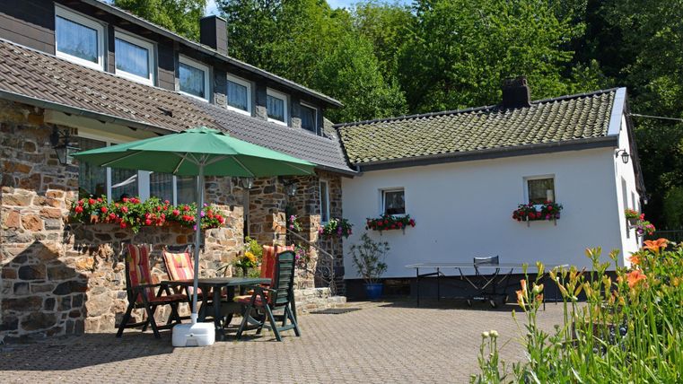 Een uitnodigend huis met een mooie buitenruimte. Er zijn tafels, stoelen en bloemen, omringd door bomen en groen.