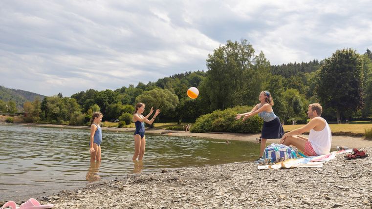 Eine Gruppe von Menschen spielt mit einem Ball am Ufer eines ruhigen Sees. Im Hintergrund sind grüne Bäume und eine entspannte Atmosphäre zu sehen.
