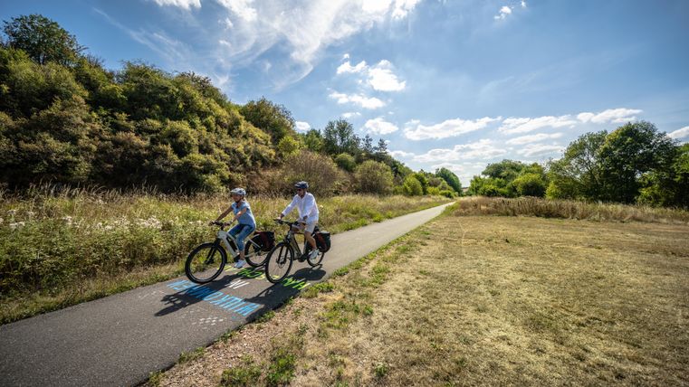 Two cyclists are riding on a quiet path through nature. In the background, there are green trees and a blue sky visible.