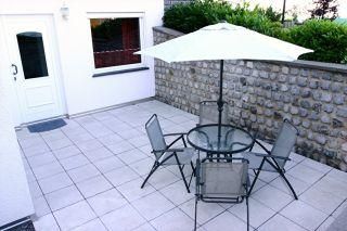 A cozy terrace with a round glass table and four chairs. A parasol provides shade and the area is surrounded by a stone wall.