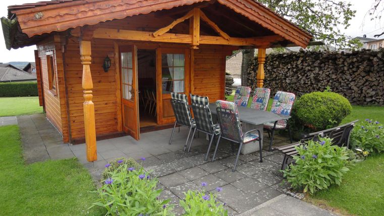 Een gezellig houten huis met een terras en een eetgedeelte buiten. Omringd door groen gras en kleurrijke bloemen.