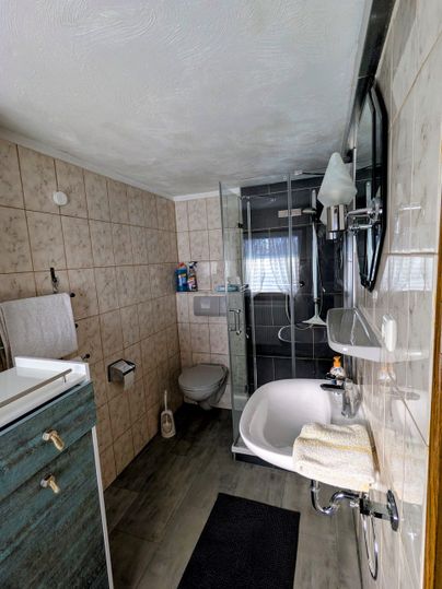 A modern bathroom with a shower, sink, and bathtub. The walls are tiled, and there is a gray mat on the floor.