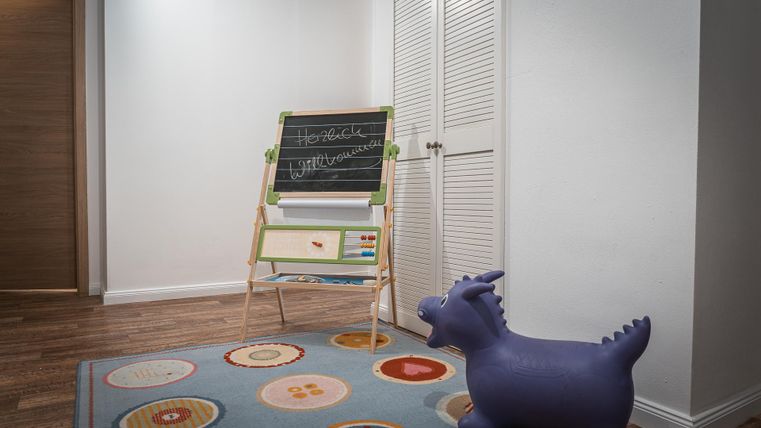 Een lichte kamer met een speelkleed en een klein, paars springspeelgoed. Aan de muur staat een klein bord om op te tekenen en te schrijven.