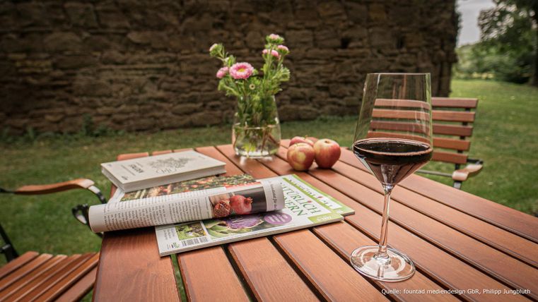 Een mooie houten tafel in de buitenlucht, versierd met een glas rode wijn, tijdschriften en een vaas met bloemen. Op de achtergrond zijn groene weilanden te zien.