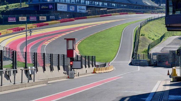 A racetrack with a long curve and a view of the grandstand. In the background, trees and a clear sky can be seen.