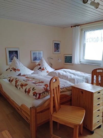 Een eenvoudige slaapkamer met twee bedden en houten stoelen. De muren zijn versierd met afbeeldingen en een raam laat veel licht binnen.