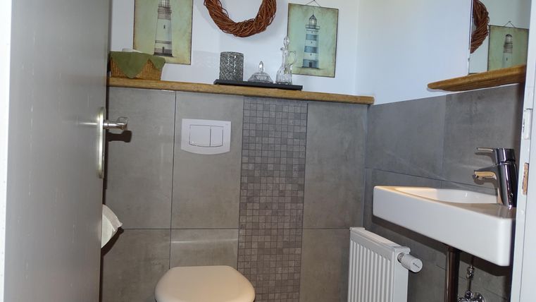 Een moderne badkamer met een wit toilet en een klein wastafel. Aan de muur hangen decoratieve schilderijen van vuurtorens en een krans.
