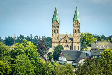 Een indrukwekkende kerk met twee spitse torens staat tussen bomen en huizen. De lucht is helder en blauw, wat de scène levendig maakt.