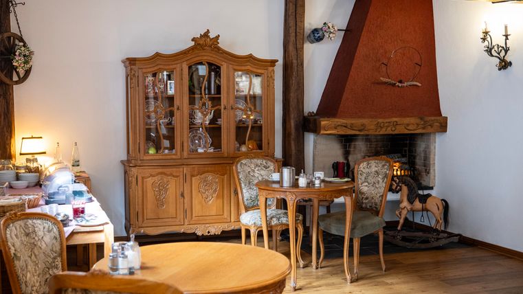 A cozy dining room with a wooden table and comfortable chairs. In the background there is a fireplace and an elegant cabinet with dishes.