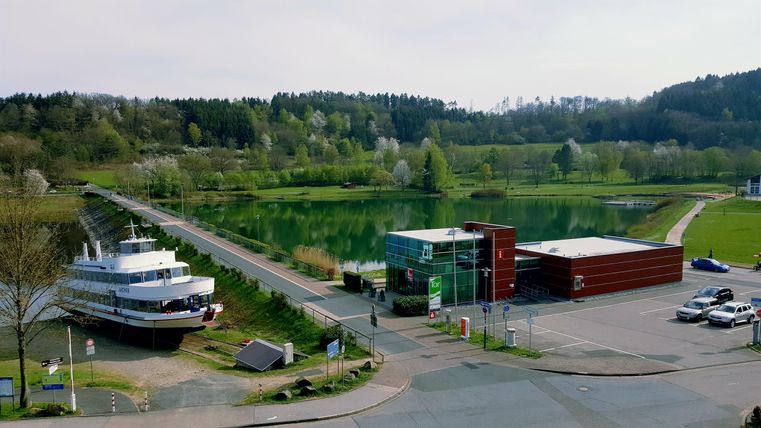 A quiet harbor with a ship and a modern building near a clear body of water.
Surrounded by green forests and meadows.