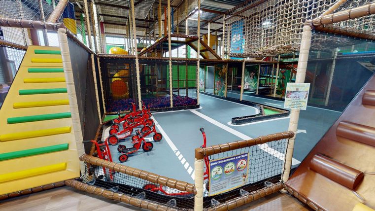 Ein bunt gestalteter Indoor-Spielplatz mit verschiedenen Spielbereichen. Es gibt eine Rutsche, Kletterelemente und ein Strafraum für Kinder mit Fahrzeugen.