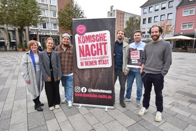 Eine Gruppe von sechs Personen steht vor einem Banner, das für eine Comedy-Veranstaltung mit dem Titel "Komische Nacht" wirbt. Die Szenerie ist urban, mit modernen Gebäuden im Hintergrund.