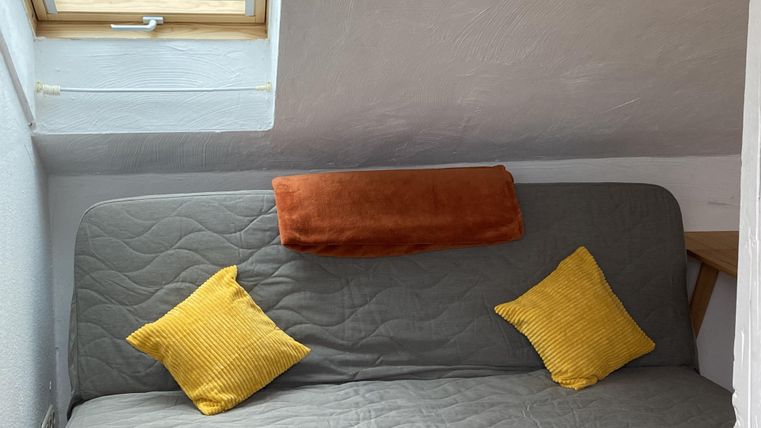 Ein gemütliches Schlafsofa mit einem grauen Bezug und drei Kissen in verschiedenen Farben. Ein Fenster sorgt für Tageslicht in dem Raum.