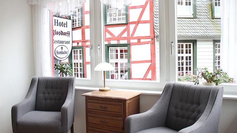 Een gezellige kamer met twee grijze stoelen en een houten tafel. Door het raam is er een mooi uitzicht op een gebouw met oranje vakwerk te zien.