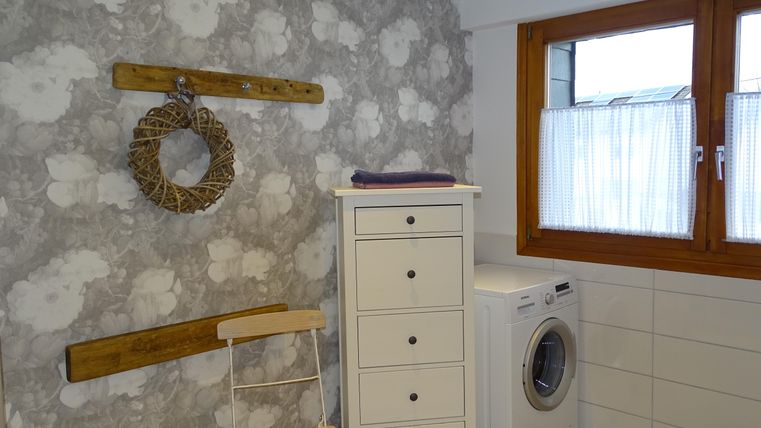 Een moderne badkamer met een lichte, gemusterde achtergrond en een groot raam. Een wasmachine staat naast een witte kast en een stoel.