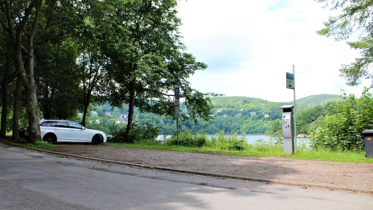 A quiet street with trees and a white car. In the background, a lake and rolling hills can be seen.