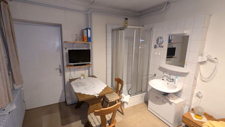 A small room with a table, two chairs, and a sink. In the corner, there is a shower and a television on the wall.