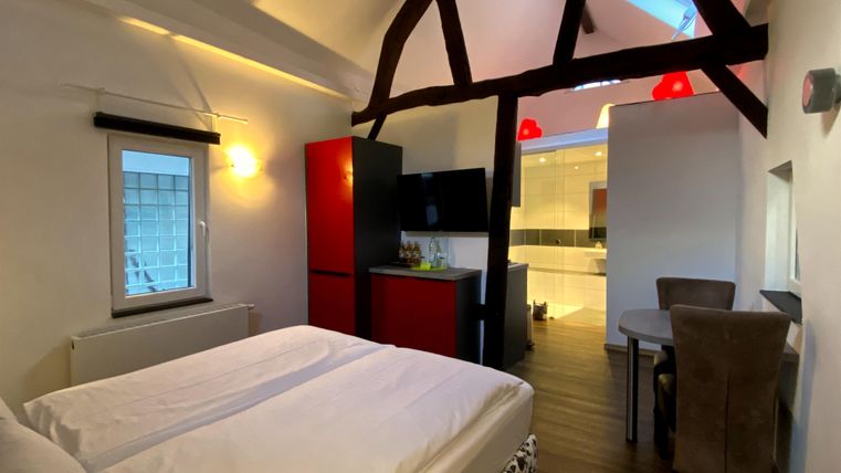 A cozy room with a double bed and wooden ceiling beams. The amenities include a refrigerator and a dining table.