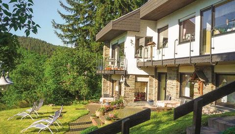 A beautiful house with large windows and a balcony amidst green spaces. In the foreground, there are loungers and flower beds.