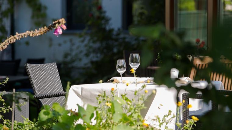A beautifully set table outdoors, surrounded by green plants and colorful flowers. On the table are two glasses and some dishes.