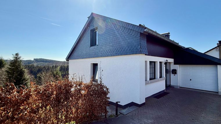Een modern huis met een blauw dak en een witte gevel. Op de achtergrond zijn bomen en een heldere lucht te zien.