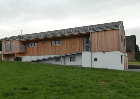 A modern, two-story house with wooden cladding. It stands on a meadow and has large windows.