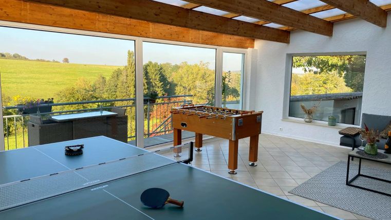 A bright room with a table tennis table and foosball, surrounded by large windows. In the background, green fields and trees are visible.