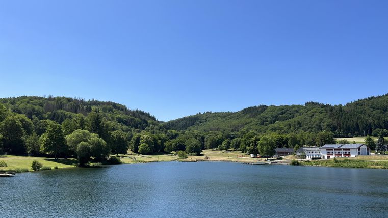 Ein ruhiger See umgeben von grünen Hügeln und Bäumen. Der klare blaue Himmel spiegelt sich im Wasser.
