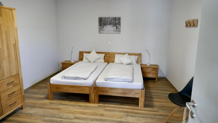 A simple, modern bedroom with two single beds and wooden furniture. The walls are in a neutral tone, and a picture of a forest hangs above the bed.