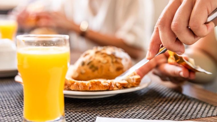 A glass of orange juice is on the table next to a buttered roll. A hand is cutting the roll, while in the background more people can be seen.