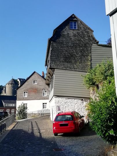 An old street with historic buildings and a red car. In the background, more buildings and a citadel can be seen.