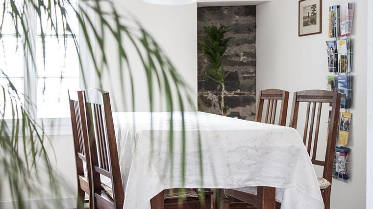 Een lichte eetruimte met een houten tafel en stoelen. Op de achtergrond zijn planten en een stenen muur te zien.