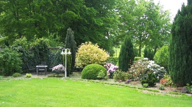 Een verzorgde tuin met kleurrijke bloemen en groene struiken. Op de achtergrond staat een bank onder schaduwgevende bomen.