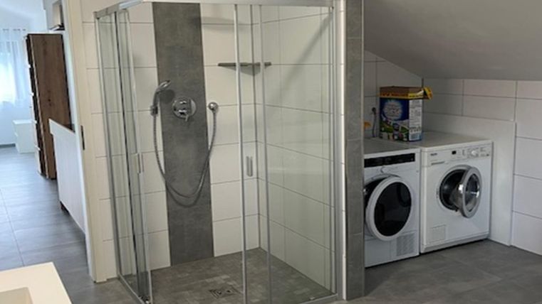 Een moderne badkamer met een glazen deur-douche en een wasmachine naast een droogmachine. De vloer is voorzien van grijze tegels en de muren zijn licht betegeld.