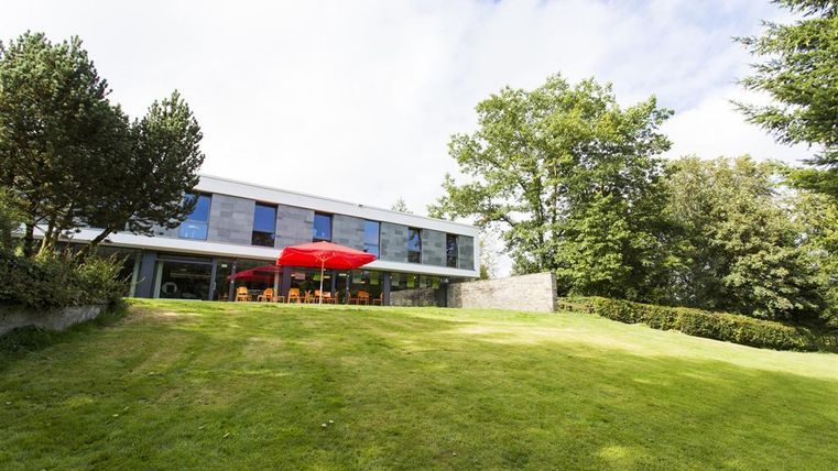 A modern house on a green meadow, surrounded by trees. In the foreground, there is a red parasol over a seating area.