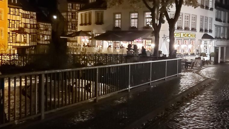 A quiet street at night with illuminated buildings and a small canal. On the edge stands a railing and the cobblestones shine from the rain.