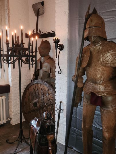 Een historische ruimte met harnassen en een kroonluchter. Te zien zijn een pantser, een schild en een houten paardenfiguur.