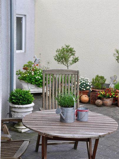 Ein kleiner, einladender Hinterhof mit einem runden Holztisch und Stühlen. Umgeben von bunten Blumen in Töpfen und grünen Pflanzen.