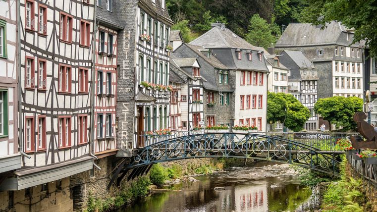 A picturesque view of a town with half-timbered houses and a small bridge over a tranquil river. Lush greenery surrounds the historic architecture.