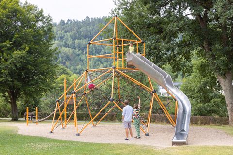 A colorful climbing frame with a slide stands in a park. In the background, trees and gentle hills can be seen.