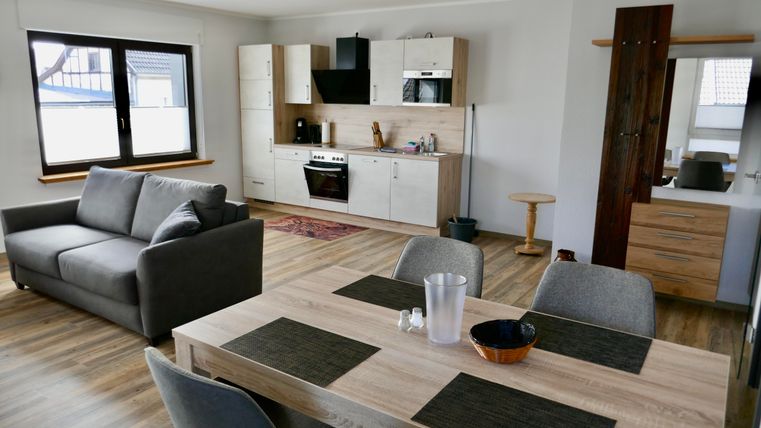 A modern apartment with an open kitchen and a cozy living area. The table is set with dishes and there is plenty of daylight coming in through the windows.