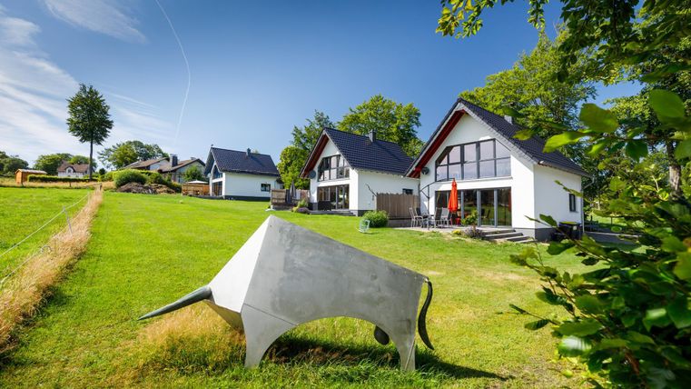 A modern house with large windows stands in the middle of a green meadow. In the foreground, there is a striking, sculptural cow made of concrete.