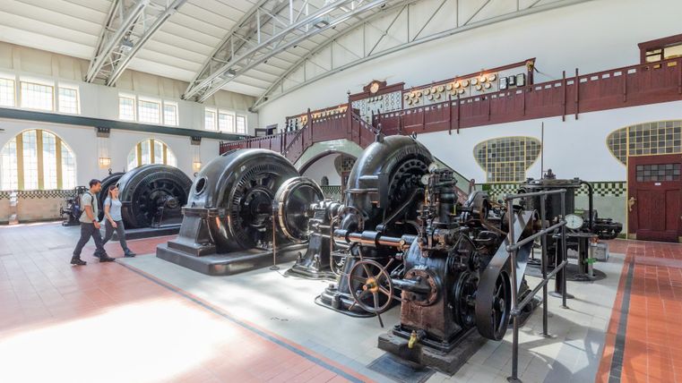 Een historisch krachtstation met grote machines en een lichte, open ruimte. Twee personen lopen door de tentoonstelling.
