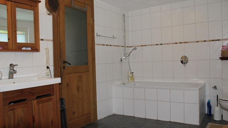 A spacious bathroom with white tiled walls and a wooden door. There is a bathtub, a shower, and a double sink.