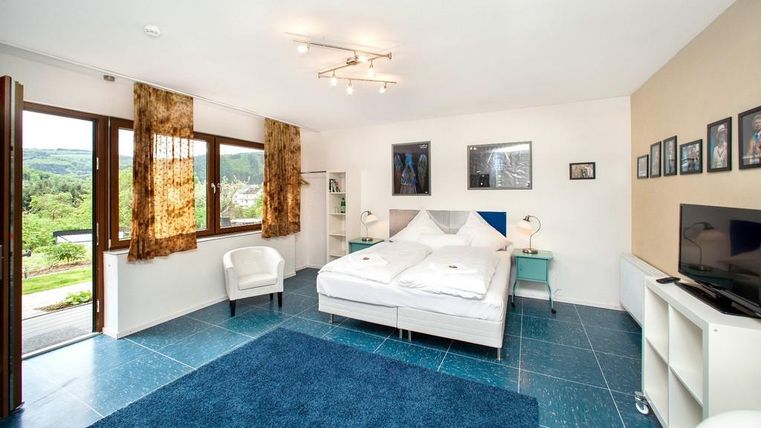 Helles und modernes Schlafzimmer mit einem großen Doppelbett und einem blauen Teppich. Fenster bieten einen Blick auf die Natur und die Einrichtung ist minimalistisch.